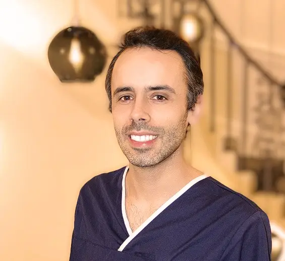 A person wearing a navy blue shirt with white trim stands indoors, smiling. In the blurred background, modern spherical pendant lights and part of a staircase are visible, reminiscent of the sophisticated ambiance found in London clinics specializing in comprehensive health screenings.