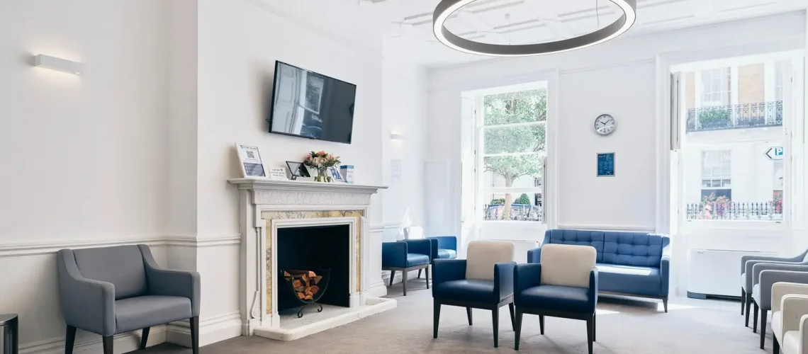 A modern waiting room in health assessment London, featuring gray and blue chairs, a fireplace, a wall-mounted TV, and large windows letting in natural light. The carpeted floor complements the decor of a clock and framed pictures, providing an inviting space for high-end health assessments.